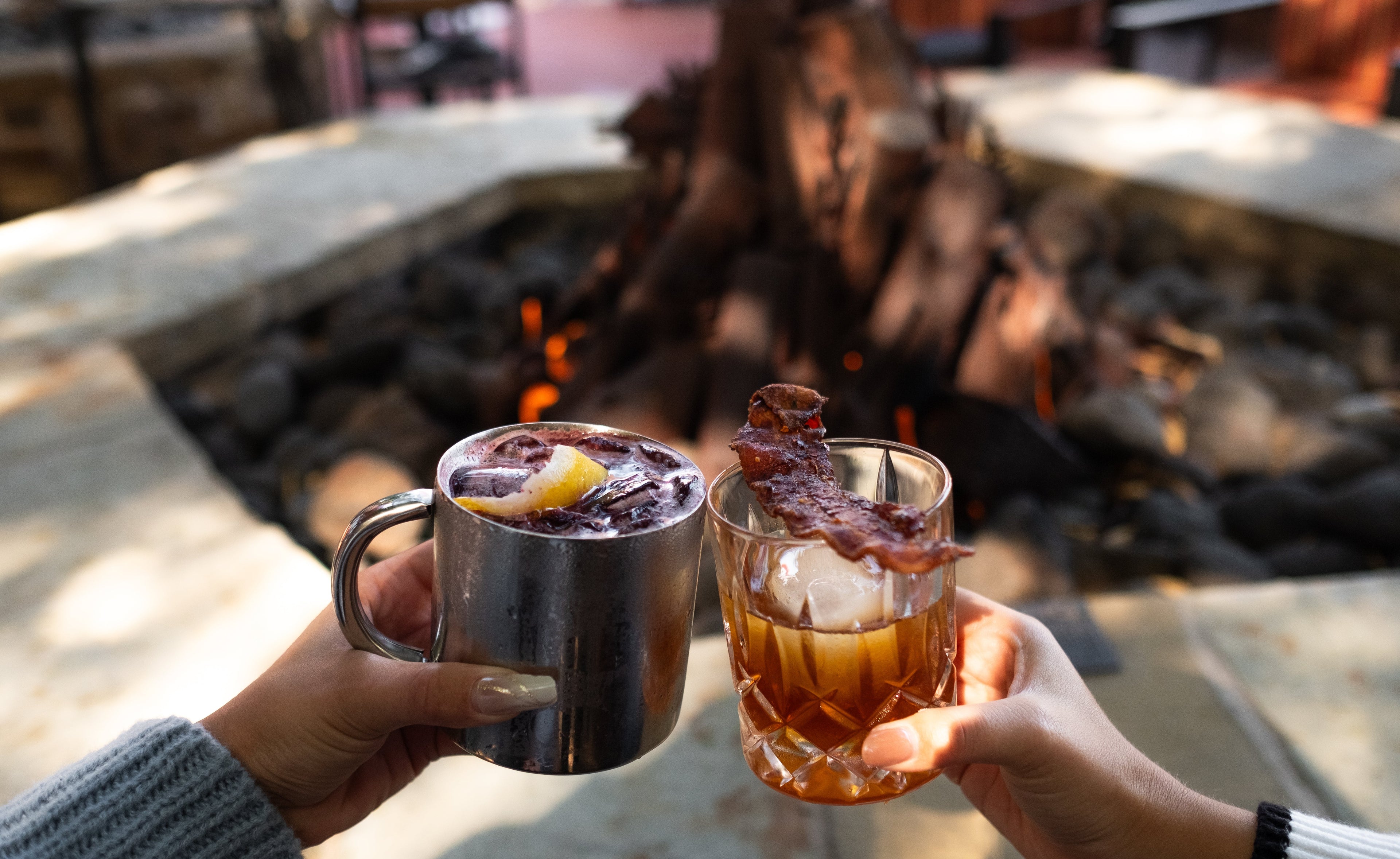Two people holding mugs with drinks by a fire pit